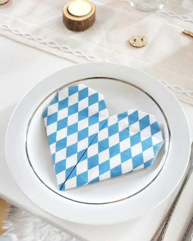 Statt kitschiger Plastikdeko kannst du deine Oktoberfest Tischdeko selber machen – rustikal, gemütlich und mit echtem Almcharakter. Viele greifen zwar schnell zu weiß-blauen Rauten und Einweg-Plastik, doch umwelttechnisch ist das kein Hit – und die Stimmung wirkt oft eher billig als festlich. DIY Oktoberfest Tischdeko basteln - Servietten zu Herz falten | Tinkerhome - Einfache DIYs für dein Zuhause